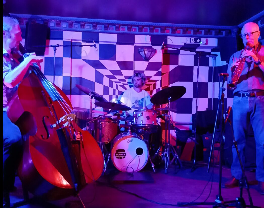 A photo of a jazz trio playing saxophone, double bass and drum kit on stage.