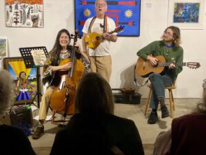 3 musicians performing in front of paintings in an art gallery in front of a seated audience.